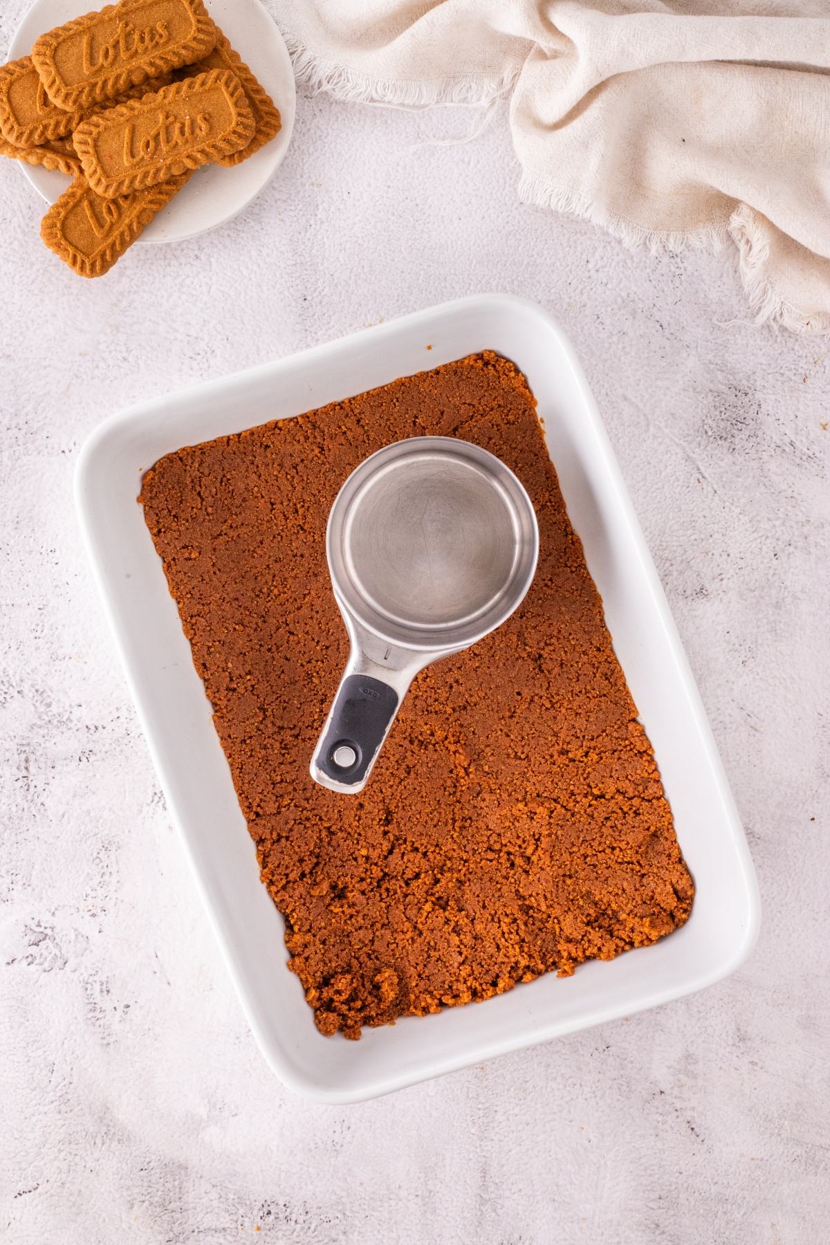 A metal cup presses cookie crumbs into a white dish; cookies are on a small plate nearby.