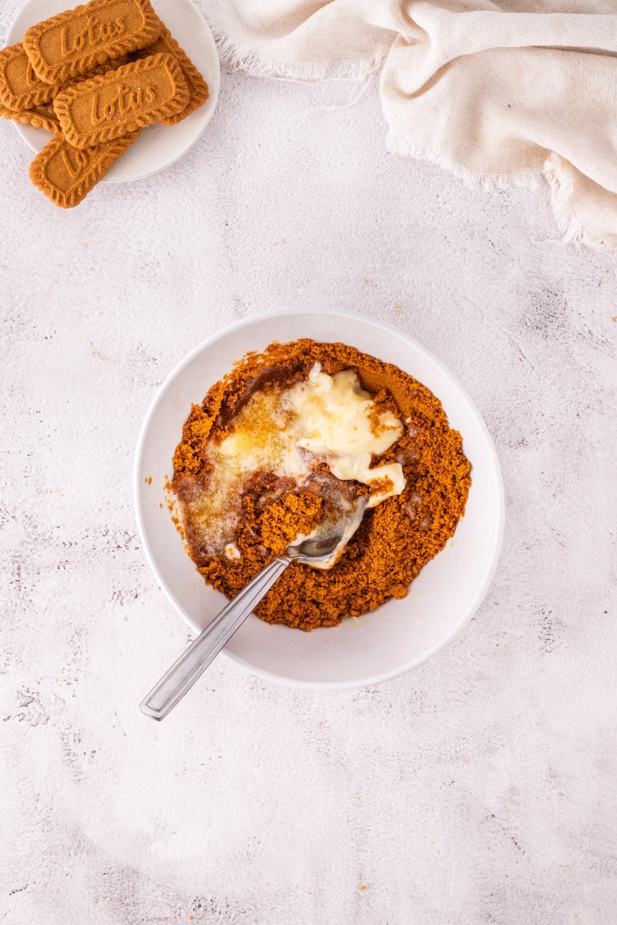 A bowl of crushed biscuits with melted butter and a spoon beside a plate of whole biscuits on a white surface.