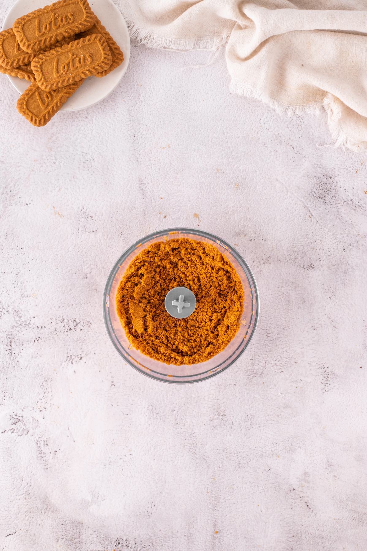 Overhead view of crushed cookies in a food processor, with whole cookies on a small plate nearby.