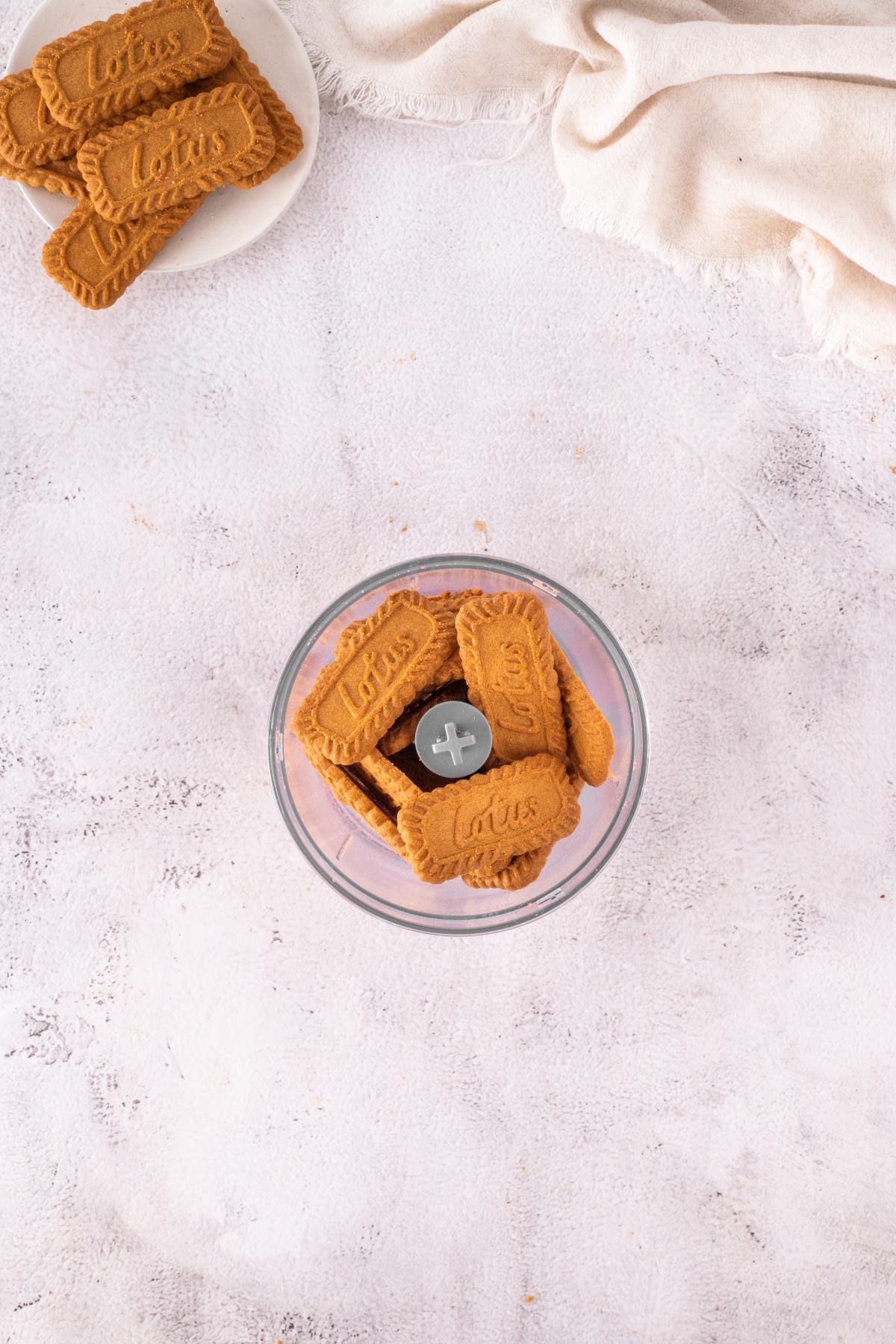 Lotus Biscoff cookies in a food processor bowl, with more cookies on a plate and a beige cloth nearby.