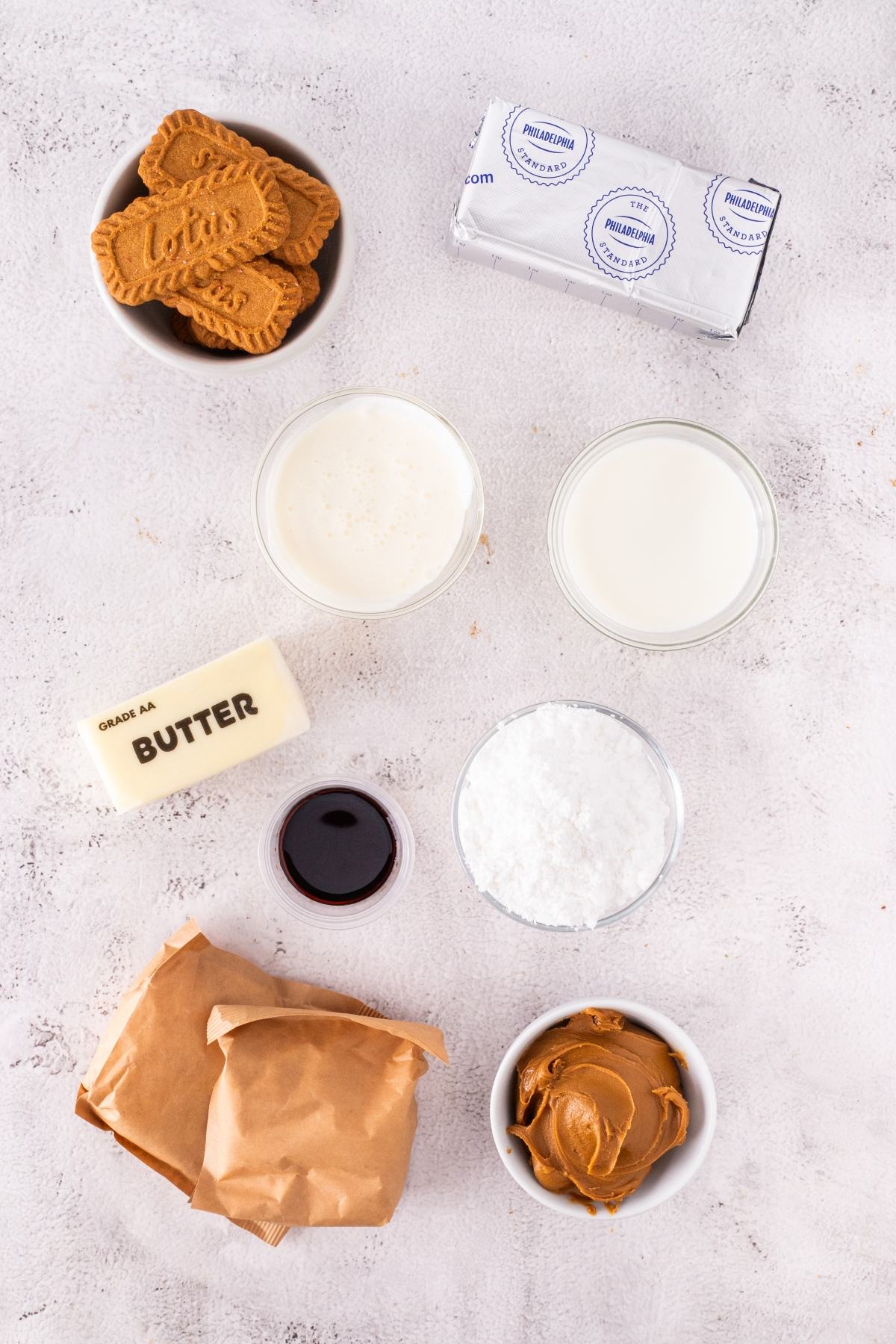 Bowls of cookies, butter, cream, milk, powdered sugar, vanilla, cream cheese, and cookie butter on a white surface.