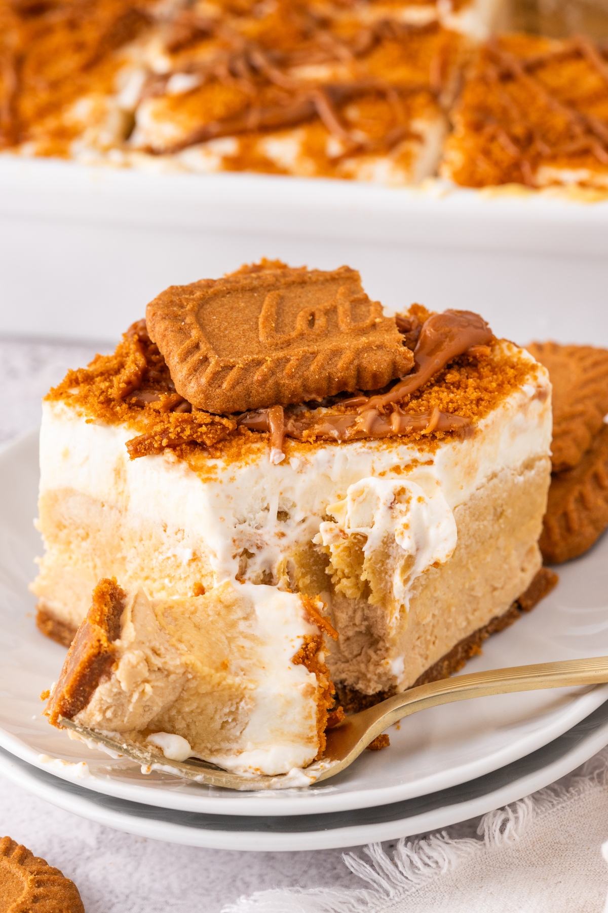 A slice of creamy biscoff lush dessert topped with a cookie and crumbs, with a forkful removed on a white plate.