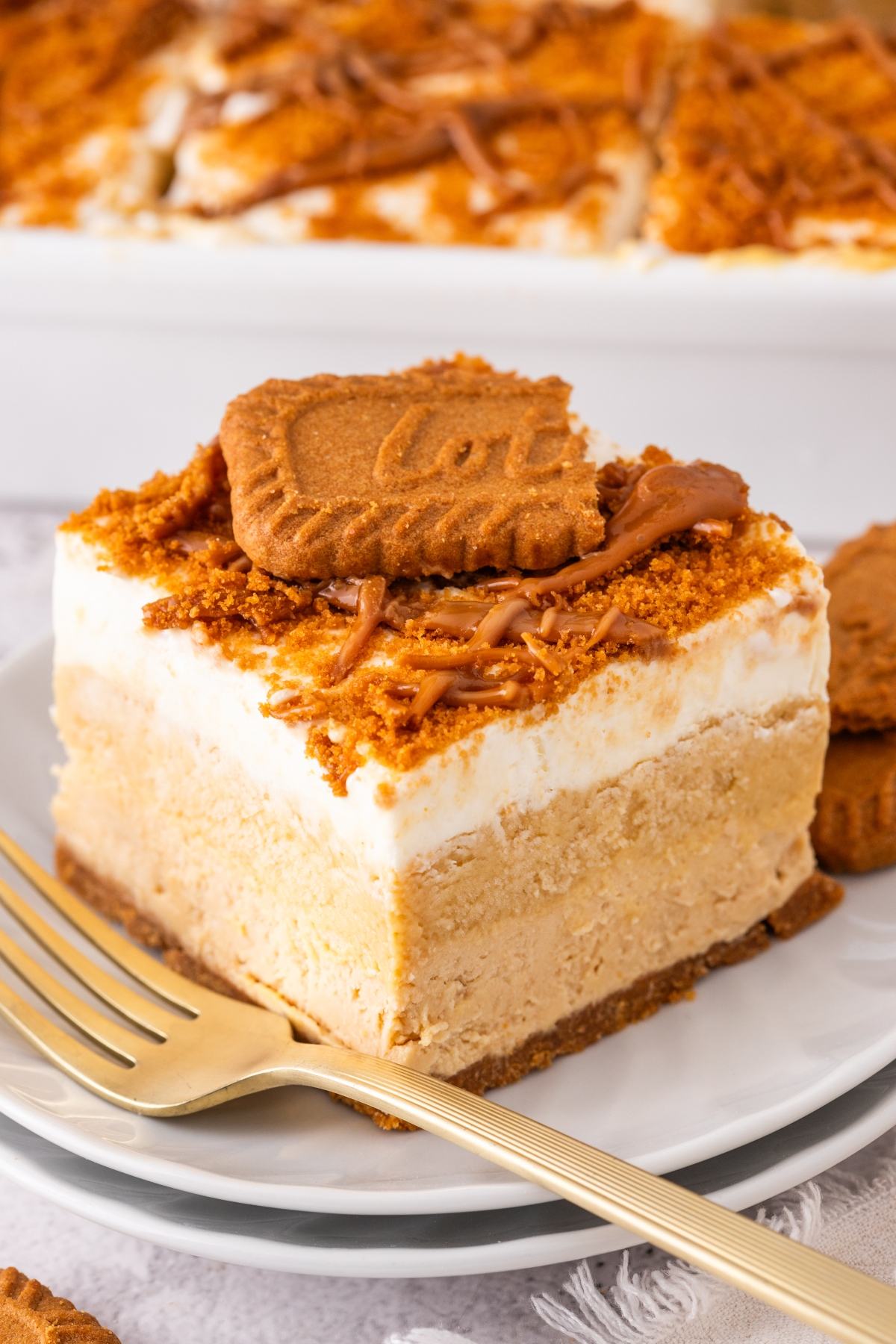A slice of layered Biscoff lush cheesecake topped with a Biscoff cookie on a plate with a gold fork.