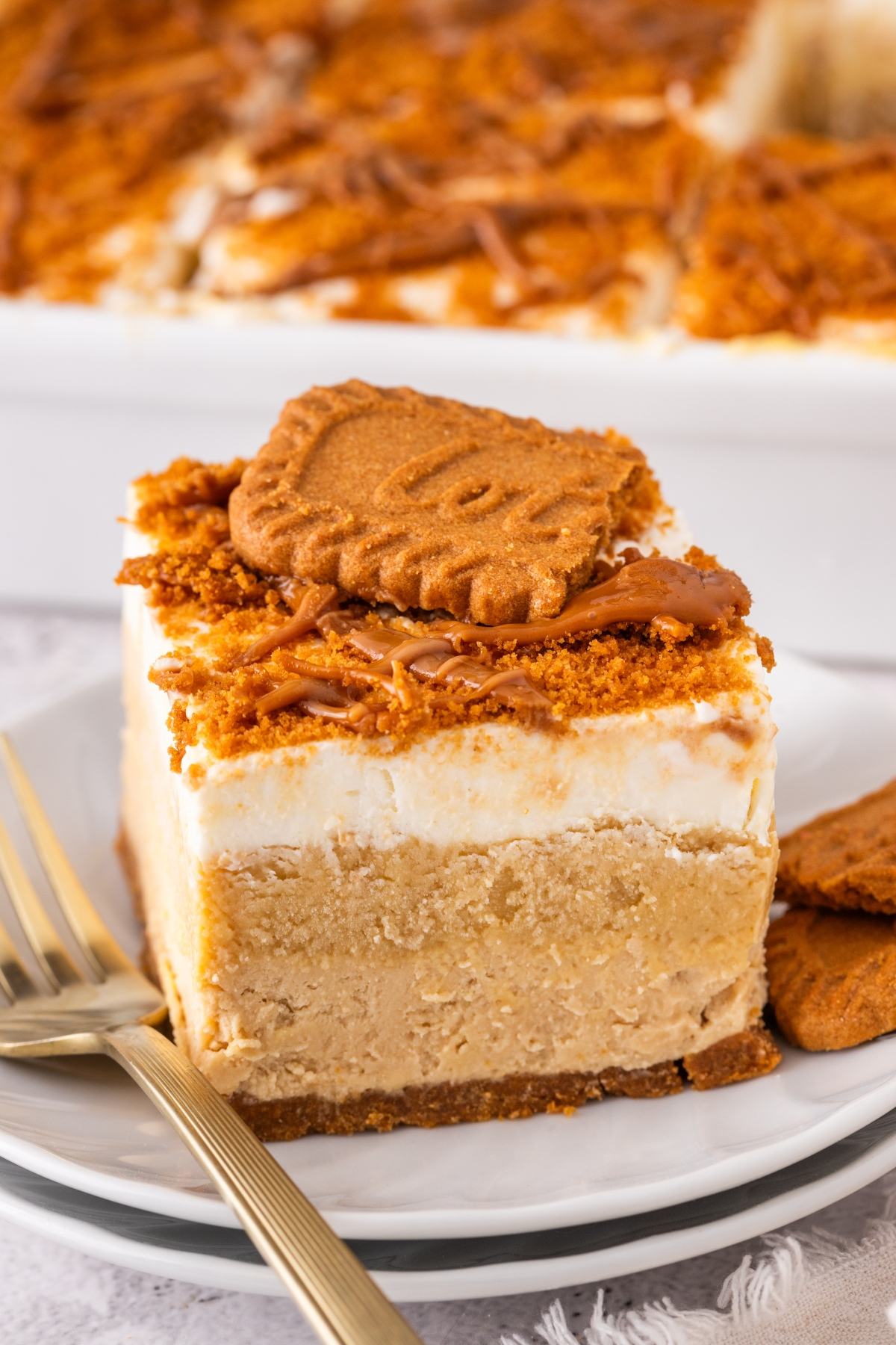 A slice of creamy biscoff lush cheesecake topped with cookie crumbles and a whole cookie, served on a white plate with a fork.