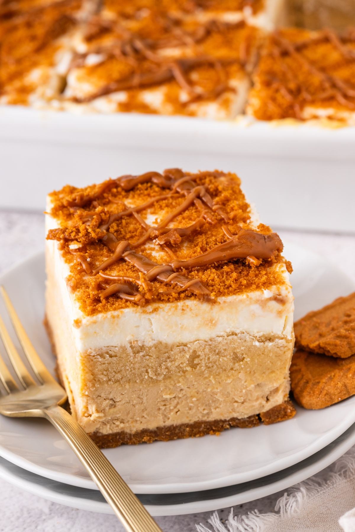 A slice of creamy layered biscoff lush dessert with a crumb topping and drizzle, served on a plate with forks and cookies.