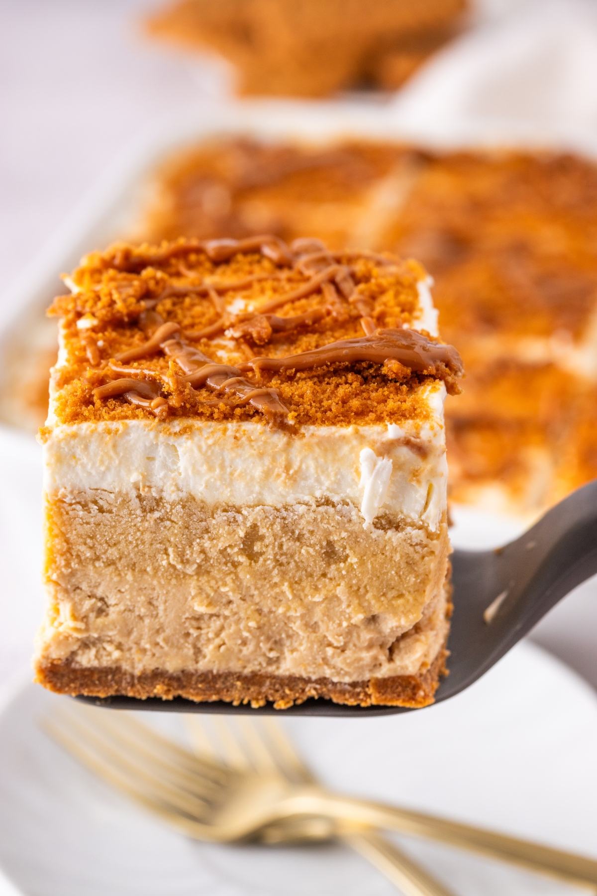 A close-up of a layered biscoff lush cheesecake bar with a crumbly topping and drizzle, held up on a spatula.