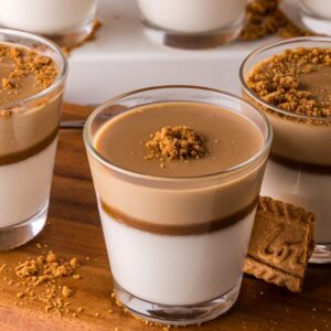 Three glasses of Biscoff Panna Cotta with caramel sauce and cookie crumbs on top, served with a Biscoff cookie on a wooden board.