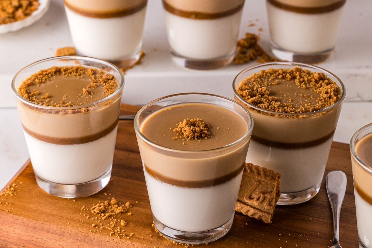 Glasses of Biscoff Panna Cotta, layered and topped with brown crumbs and a cookie, are arranged on a wooden surface.