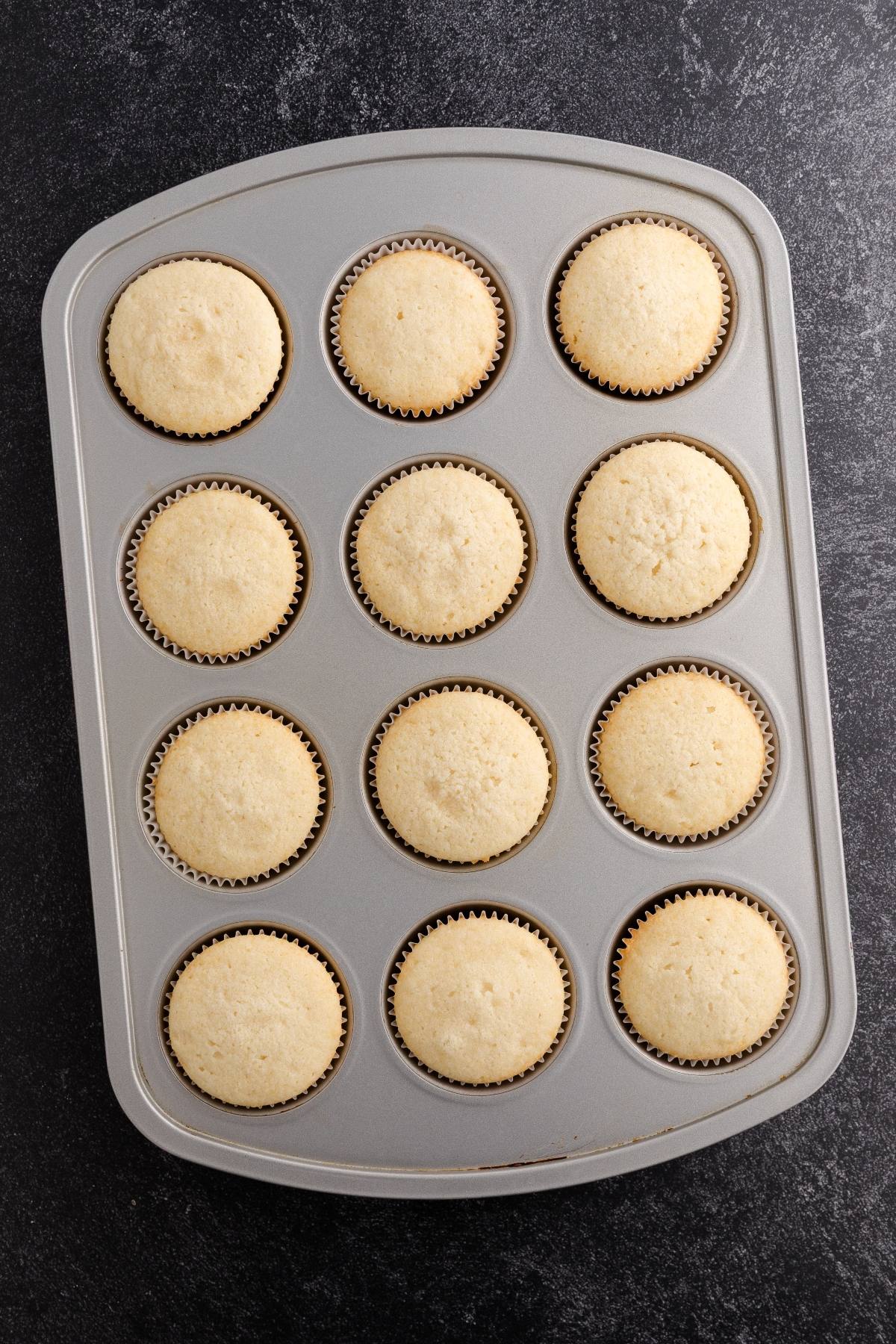 A muffin tin with twelve plain vanilla cupcakes on a dark countertop.