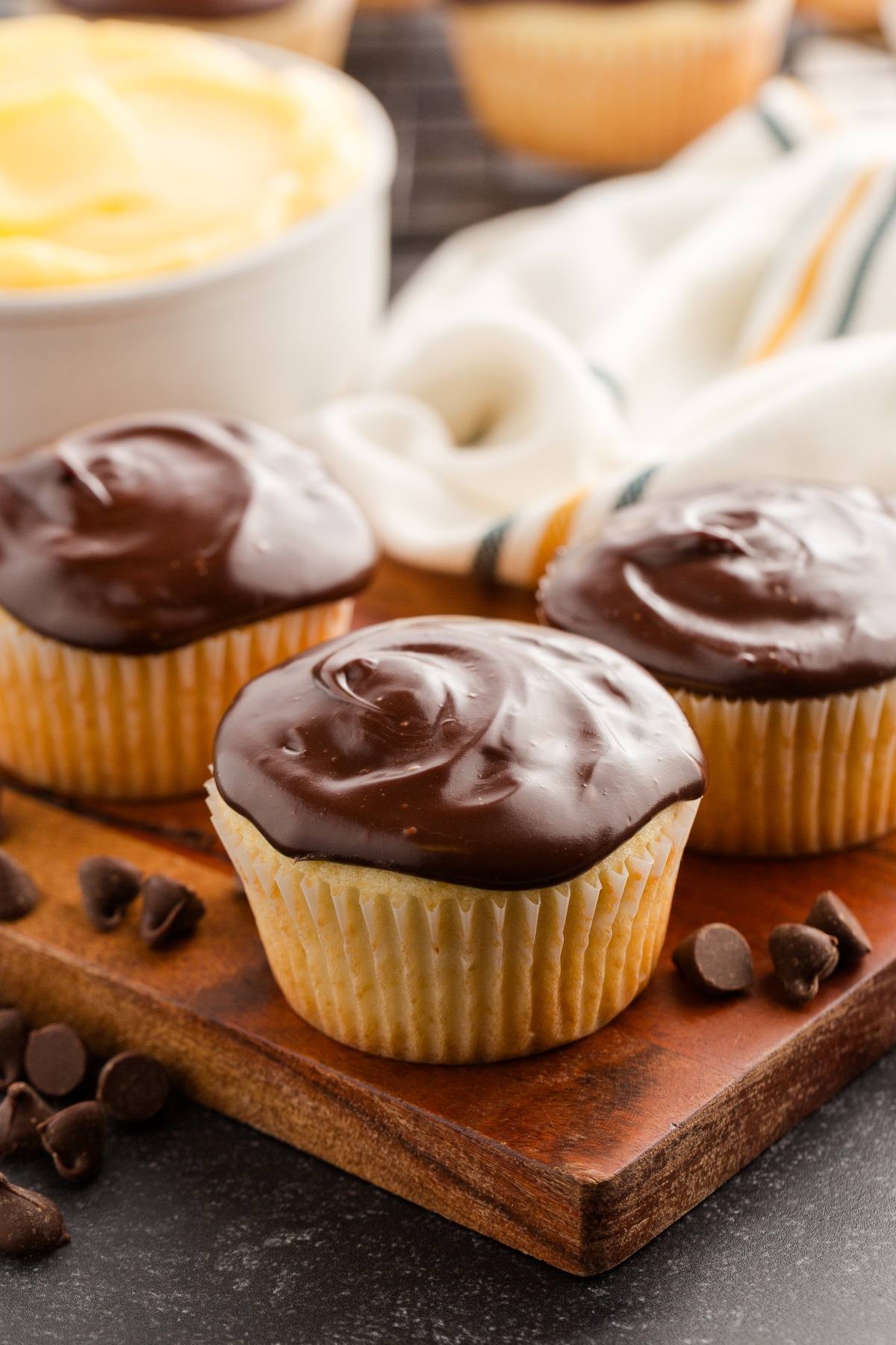 Three vanilla boston cream cupcakes with chocolate frosting on a wooden board, with chocolate chips and a cloth nearby.