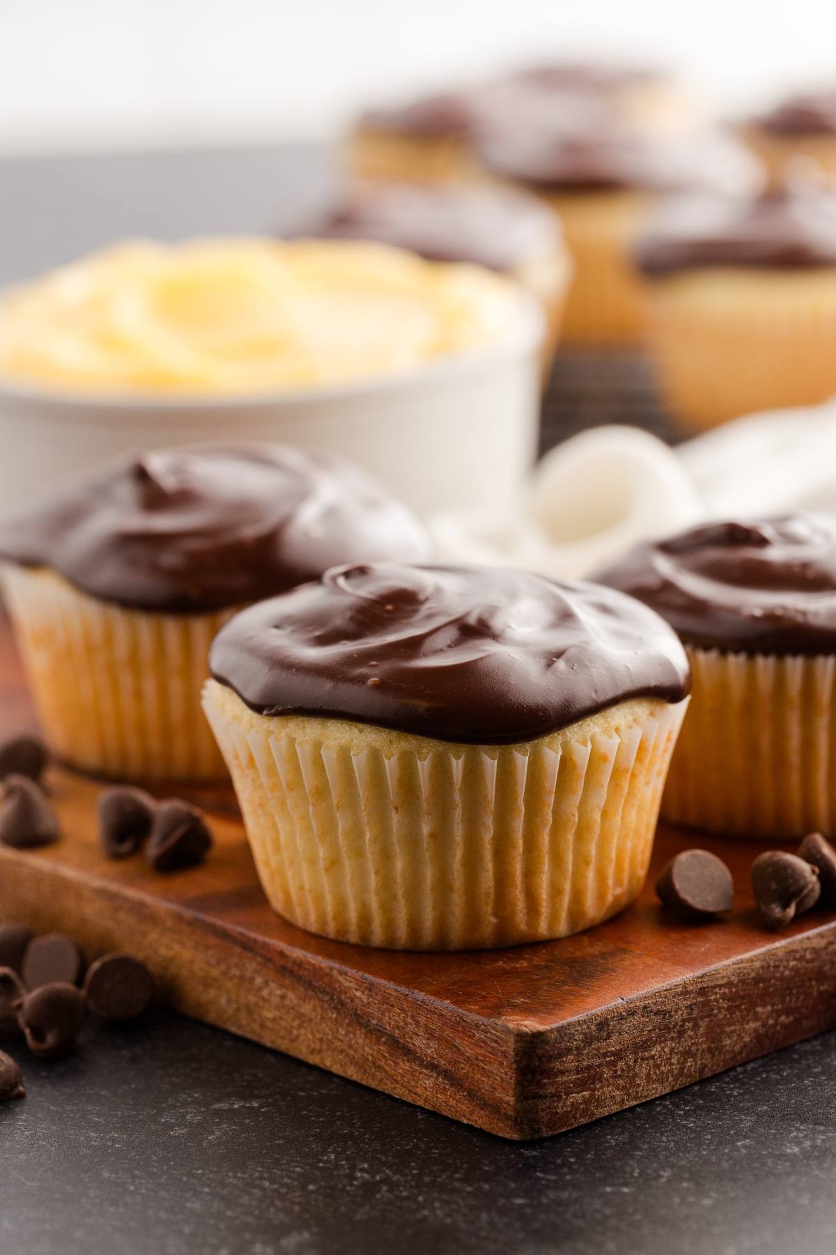 Vanilla boston cream cupcakes topped with chocolate frosting, surrounded by chocolate chips on a wooden board.