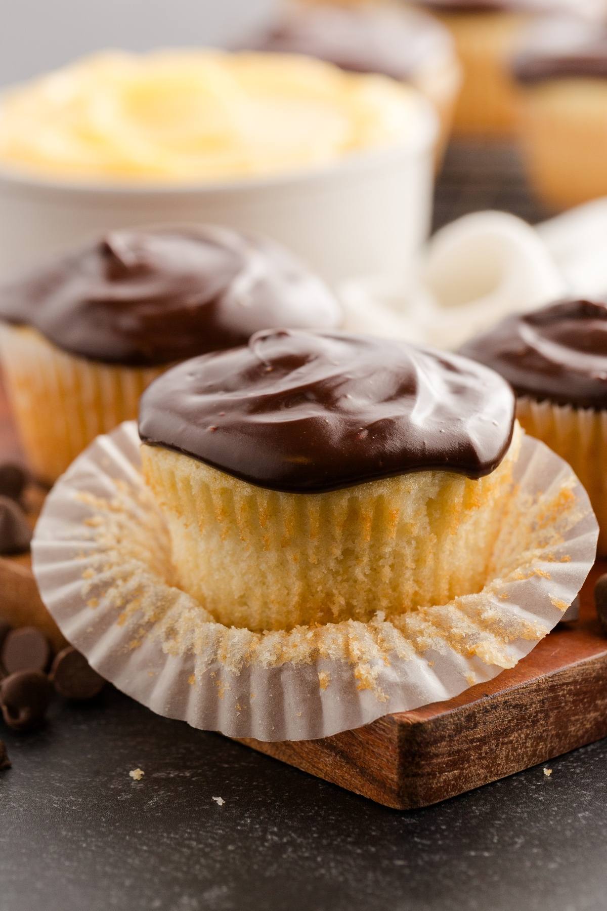 A vanilla boston cream cupcake with chocolate frosting and an open wrapper, with more cupcakes in the background.