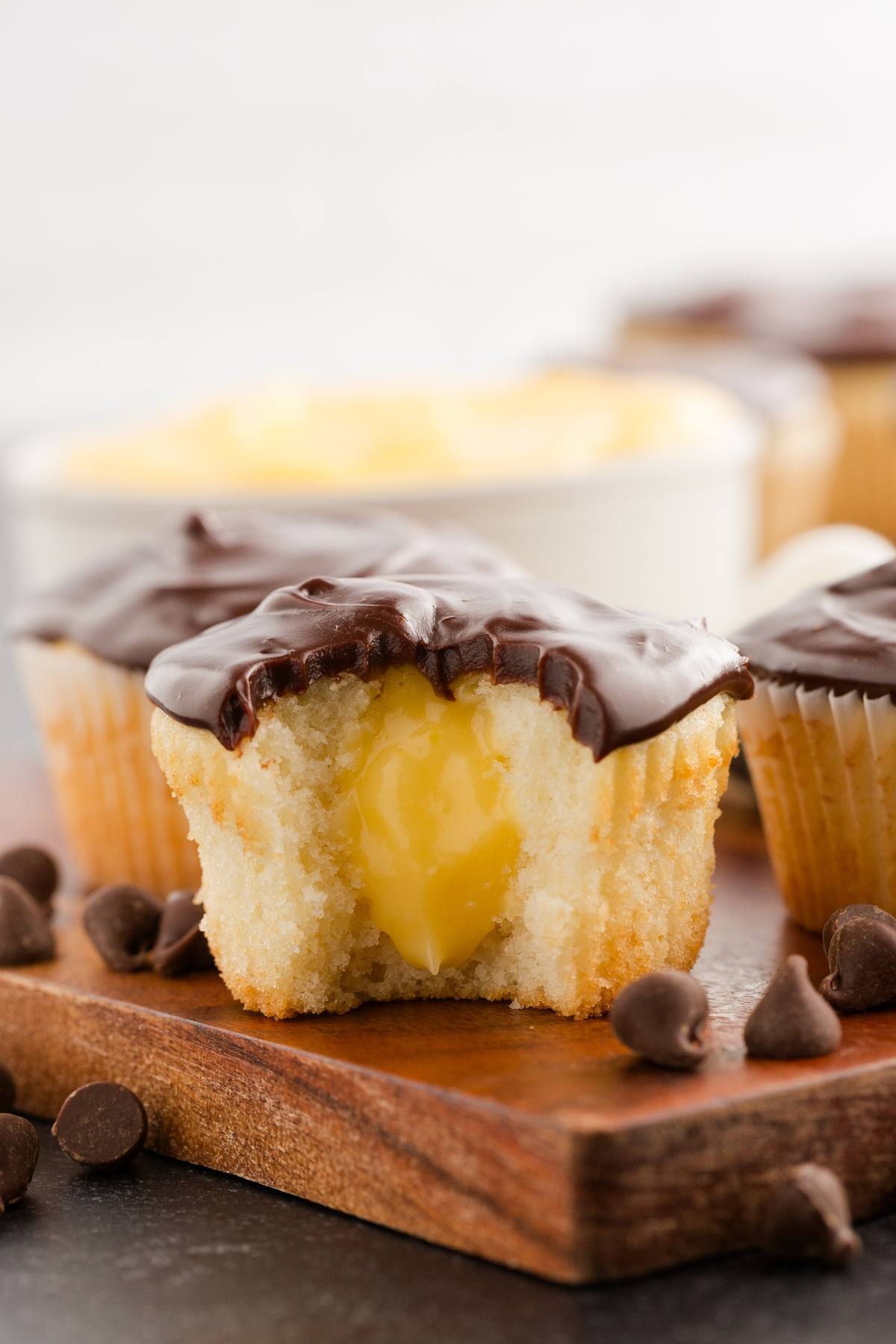 A vanilla boston cream cupcake with chocolate icing and a creamy yellow filling sits on a wooden board with chocolate chips.