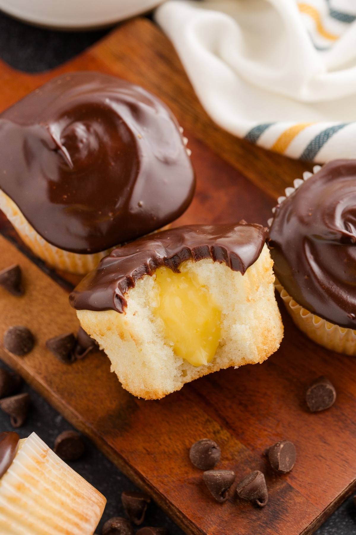 Vanilla Boston cream cupcake with chocolate frosting, cut open to reveal creamy yellow filling, on a wooden board.
