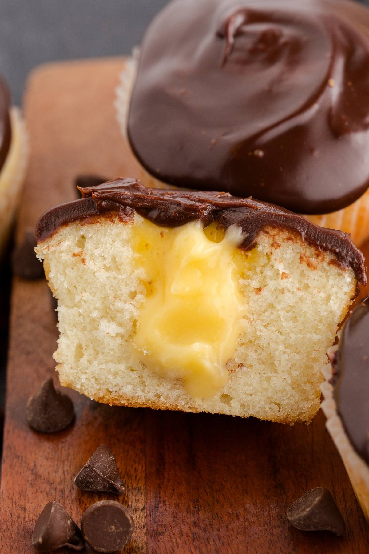 A Boston cream  cupcake with creamy yellow filling and chocolate frosting, cut in half on a wooden board with chocolate chips.