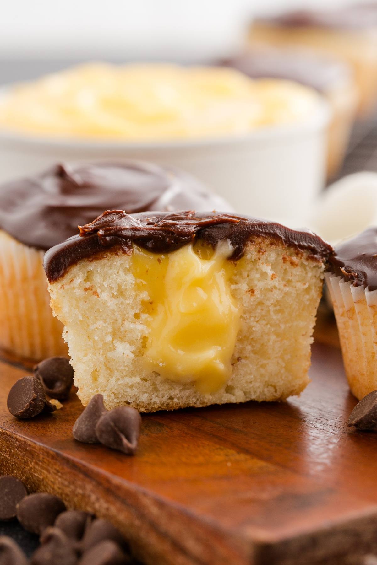 A Boston cream  cupcake with chocolate frosting and yellow cream filling, cut in half on a wooden board with chocolate chips.