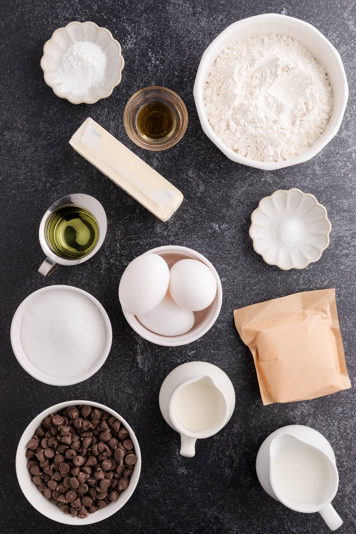 Baking ingredients on a dark surface: flour, eggs, sugar, butter, chocolate chips, milk, oil, and bowls.