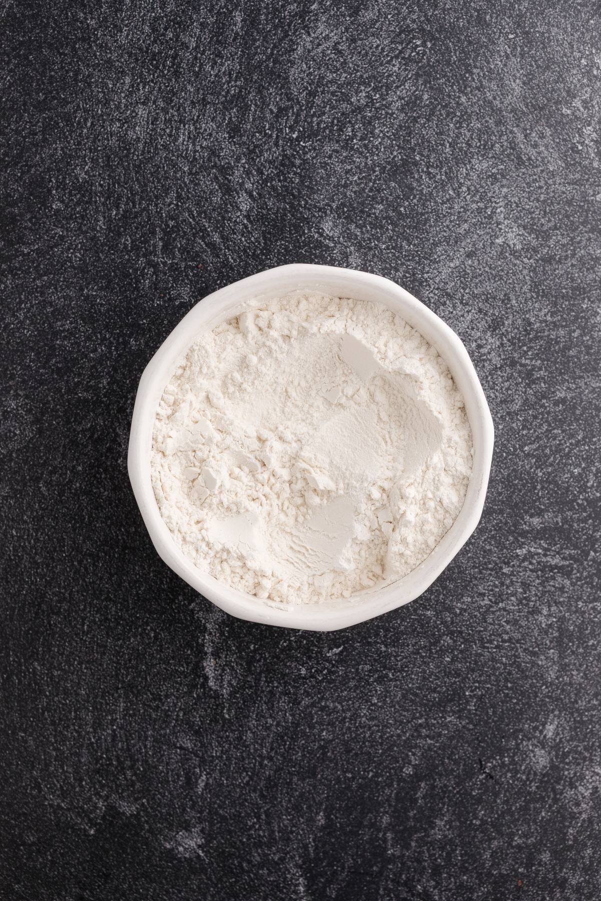A white bowl filled with flour on a dark, textured surface.