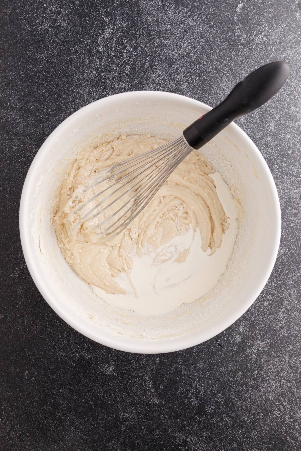 A whisk in a white bowl mixing light-colored batter on a dark countertop.