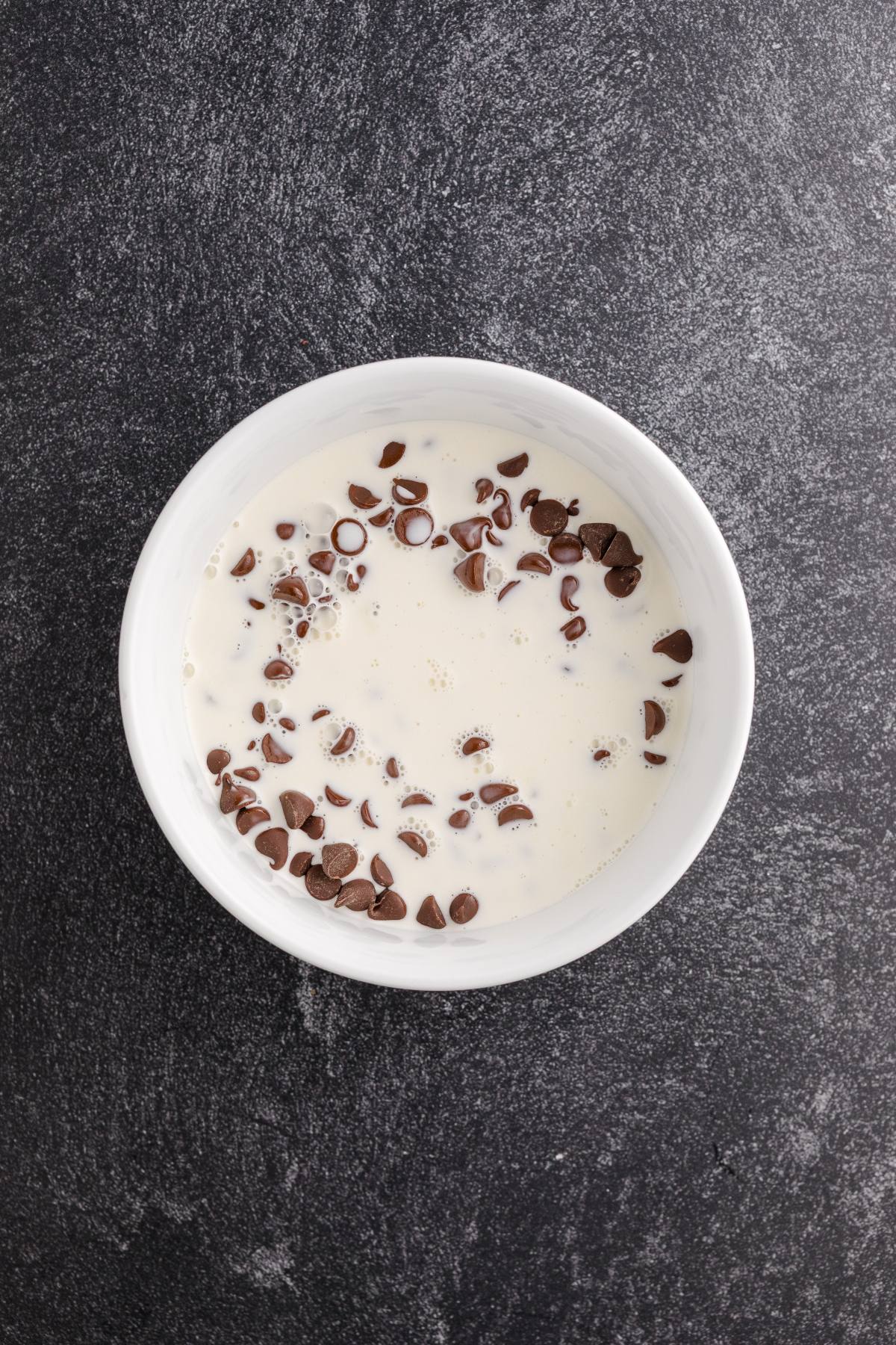 A white bowl filled with milk and chocolate chips, on a dark textured surface.