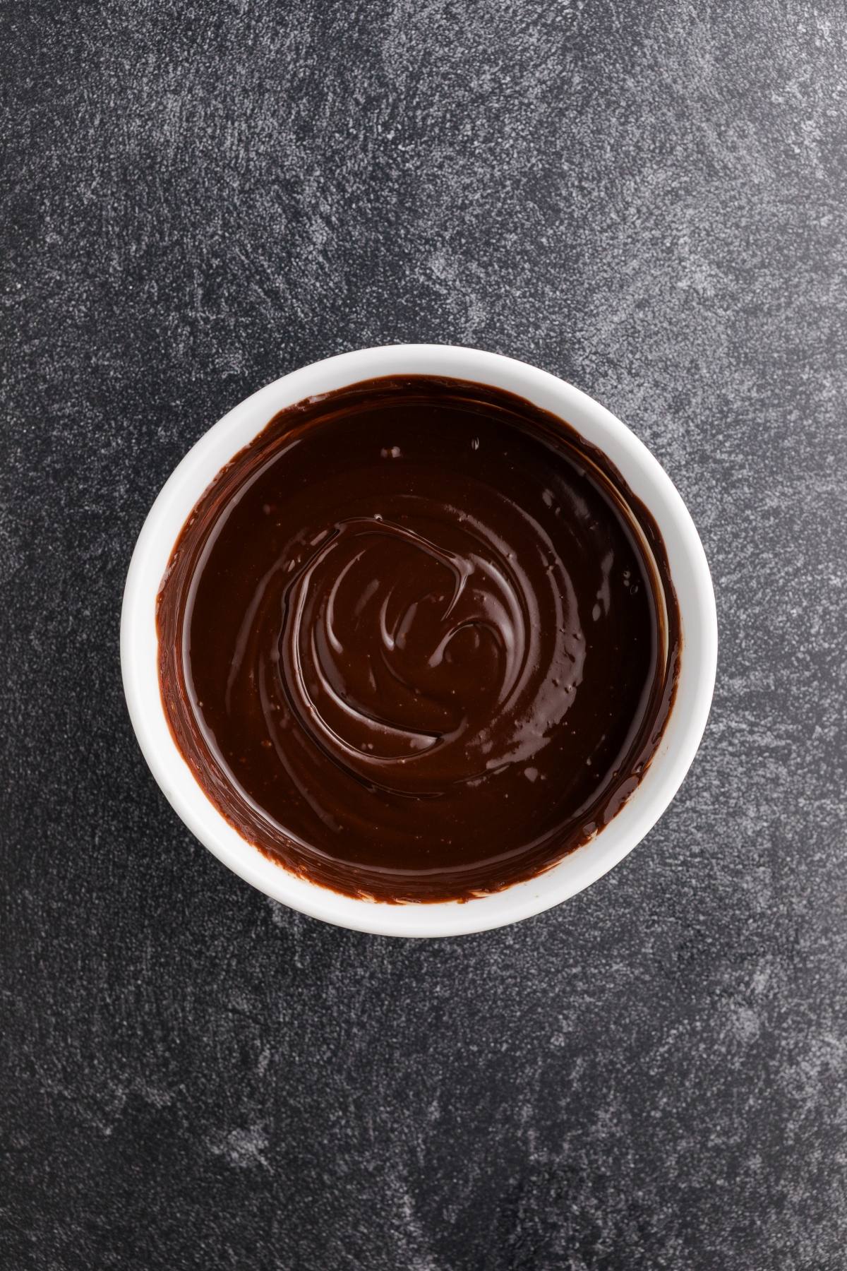 A white bowl filled with smooth melted chocolate sits on a dark, textured surface.