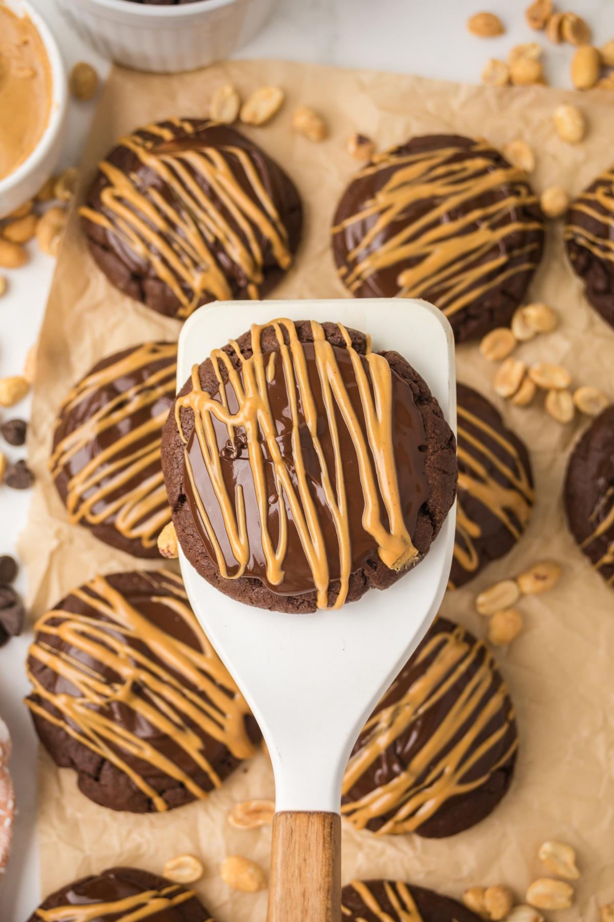 A spatula holds a chocolate cookie drizzled with peanut butter, surrounded by similar cookies and peanuts.