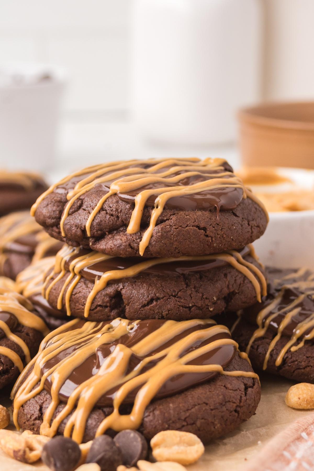 Three stacked chocolate cookies drizzled with peanut butter, surrounded by more cookies, nuts, and chocolate chips.