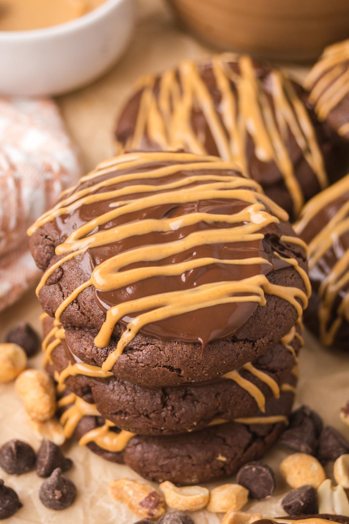 Stacked chocolate cookies drizzled with peanut butter, surrounded by peanuts and chocolate chips.