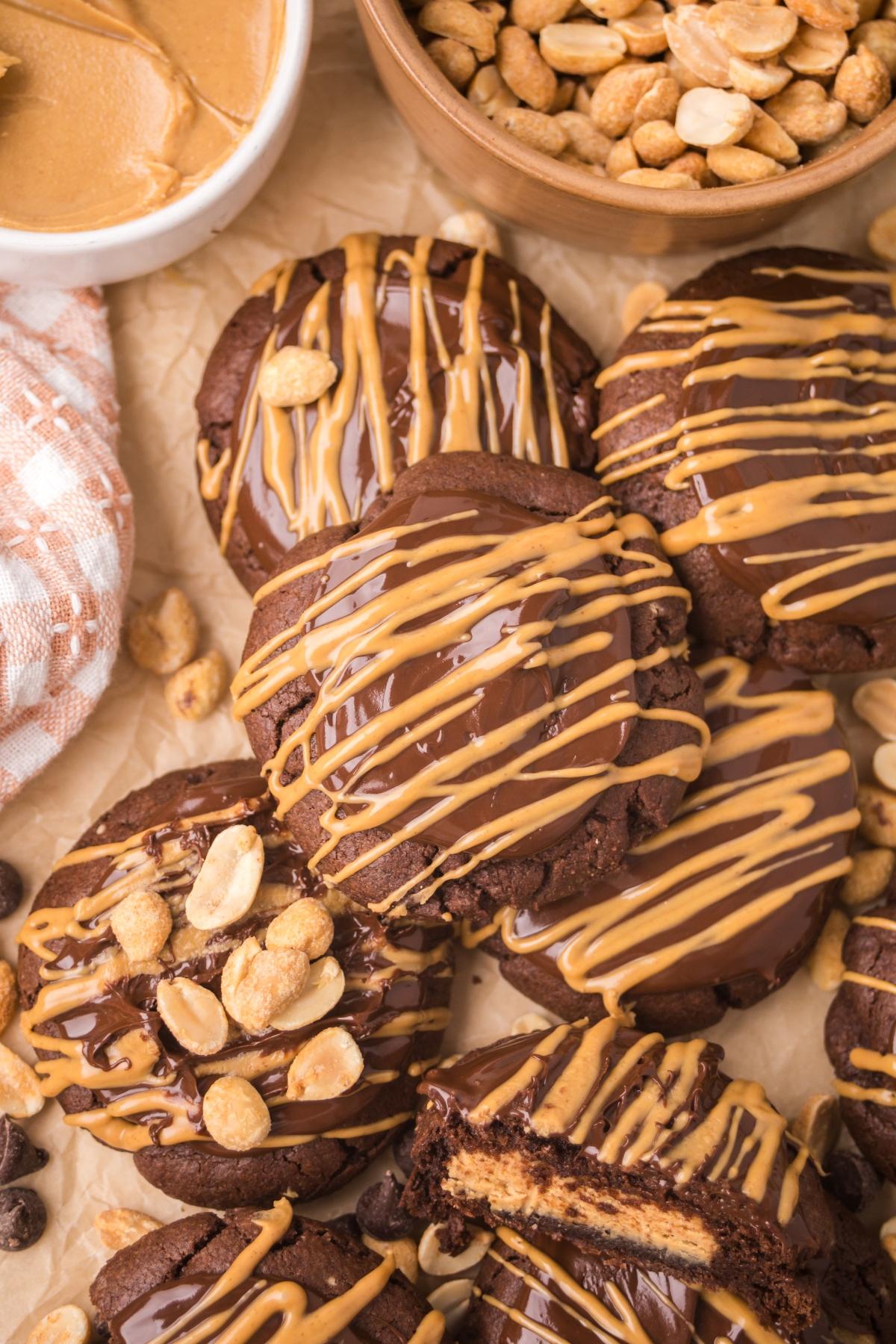 Chocolate cookies drizzled with peanut butter, surrounded by peanuts and a bowl of creamy peanut butter.