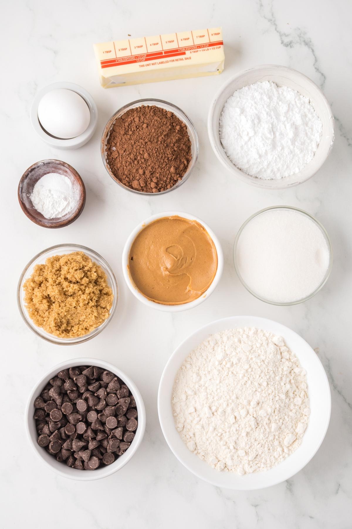 Bowls of baking ingredients, including flour, sugar, cocoa, chocolate chips, peanut butter, egg, and butter.
