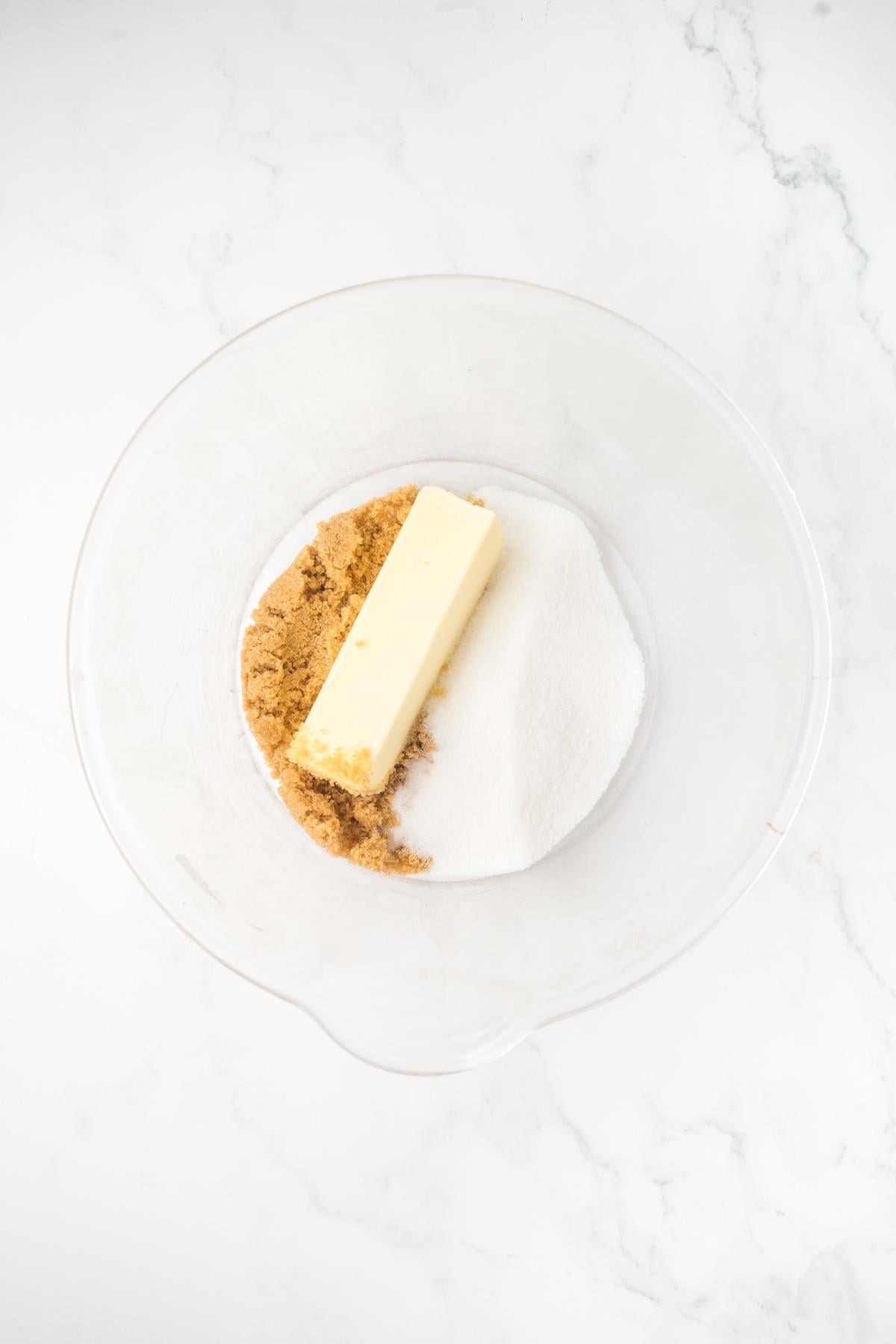 A glass bowl with butter, brown sugar, and white sugar on a white marble surface.