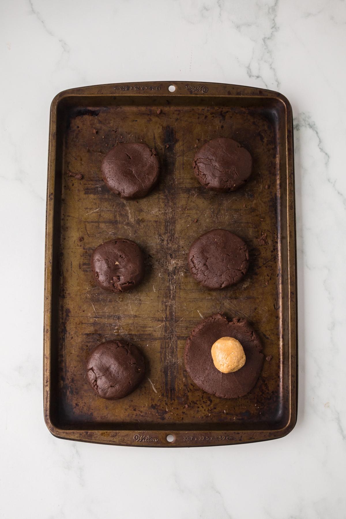 A baking sheet with six chocolate cookie dough balls, one with a peanut butter center, on a white surface.
