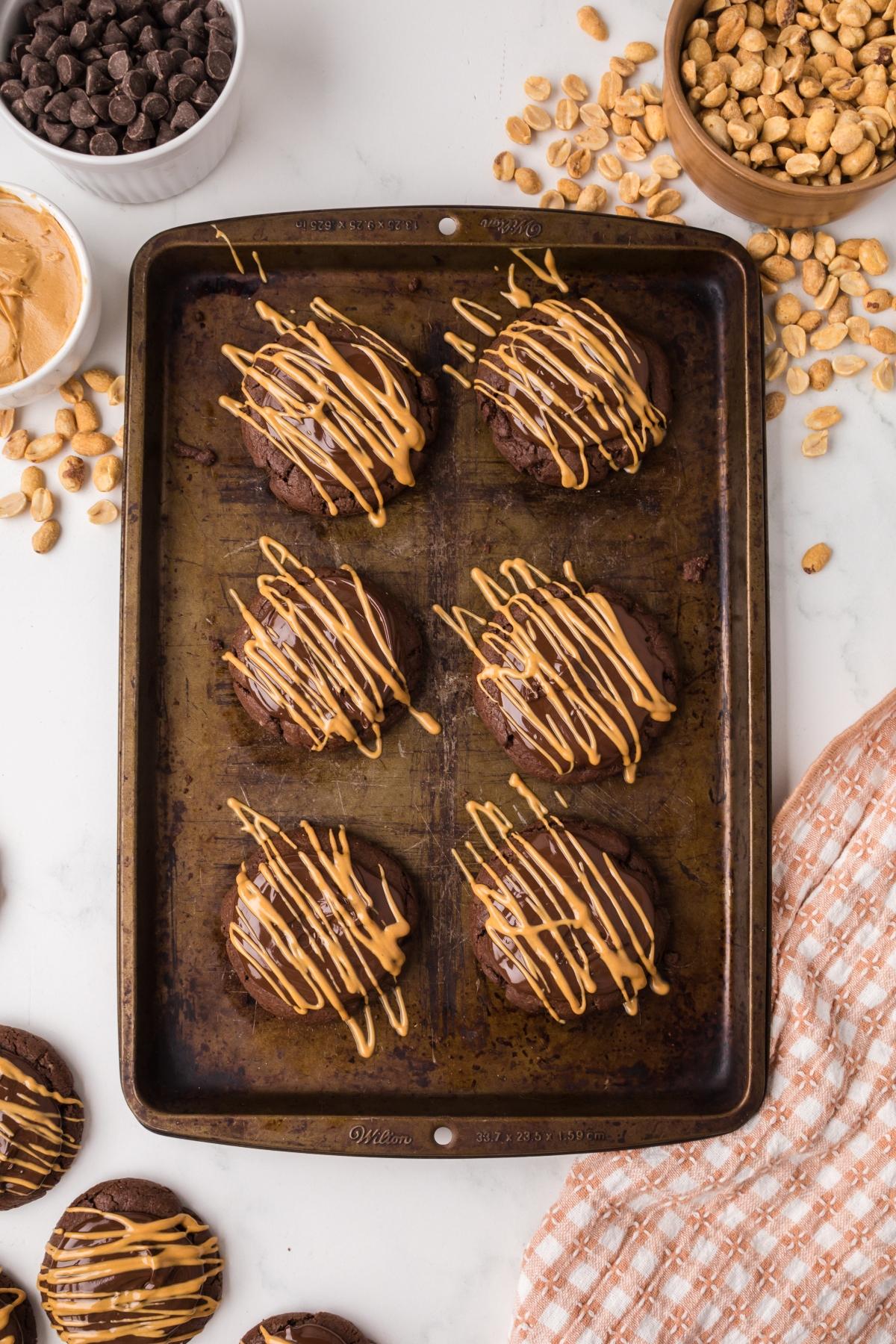 Six chocolate cookies drizzled with peanut butter on a baking tray, surrounded by peanuts and chocolate chips.