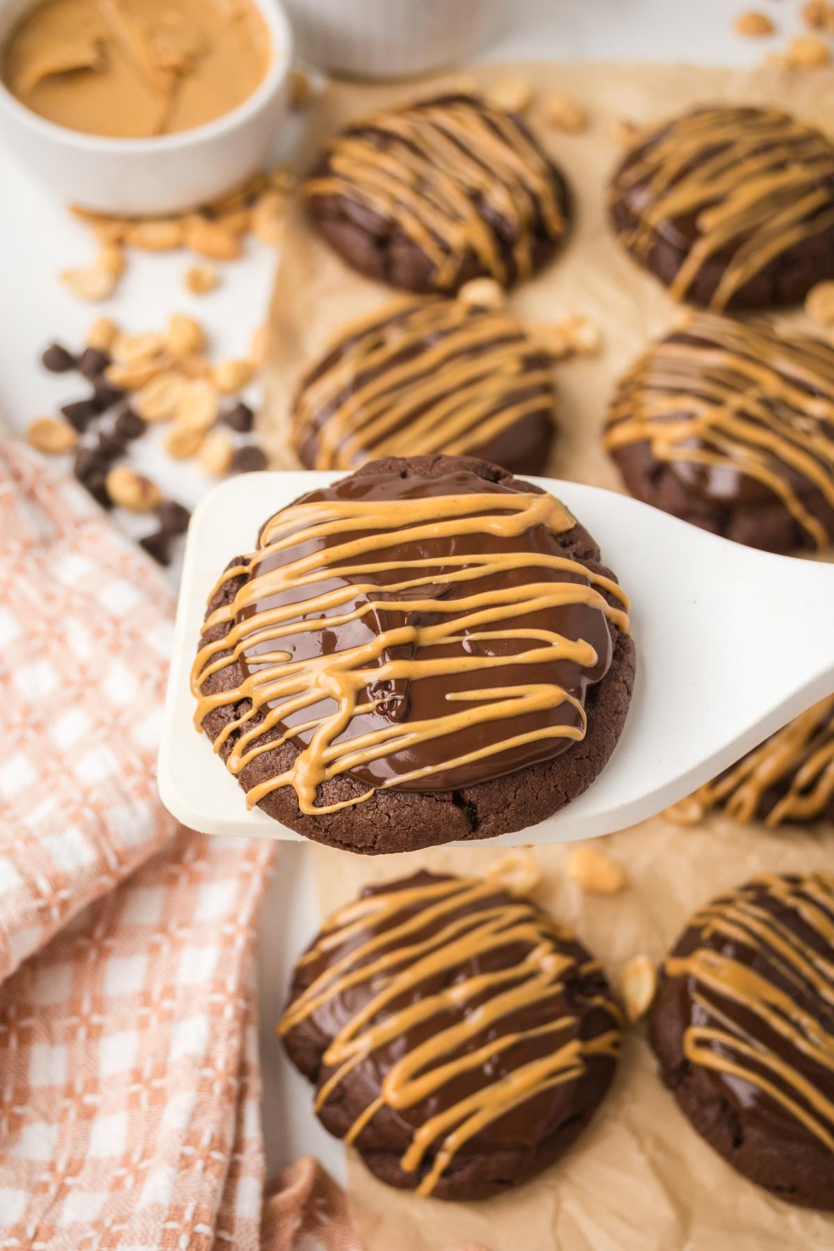A spatula holds a chocolate cookie drizzled with peanut butter, with more cookies on parchment below.