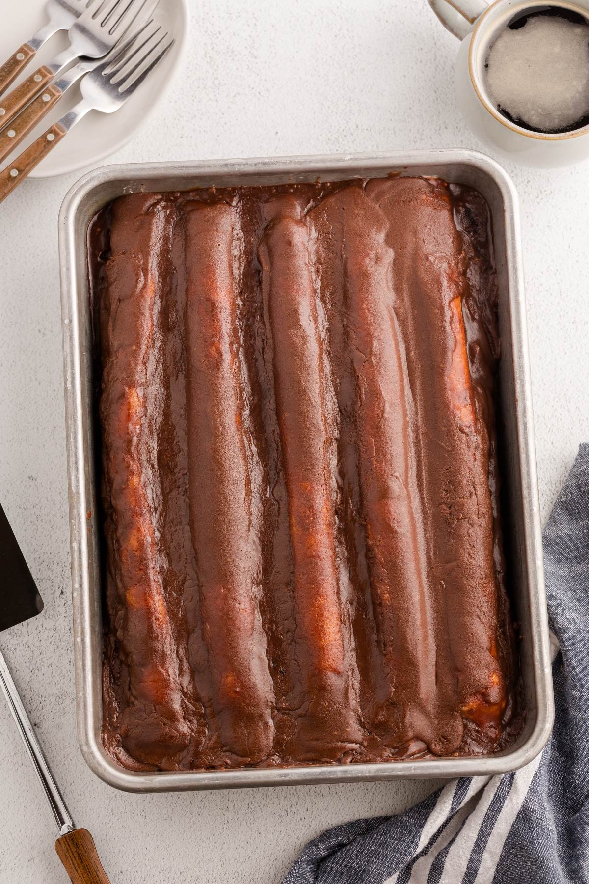 A pan of chocolate-frosted cake rolls sits on a white table beside a mug, forks, and a striped towel.