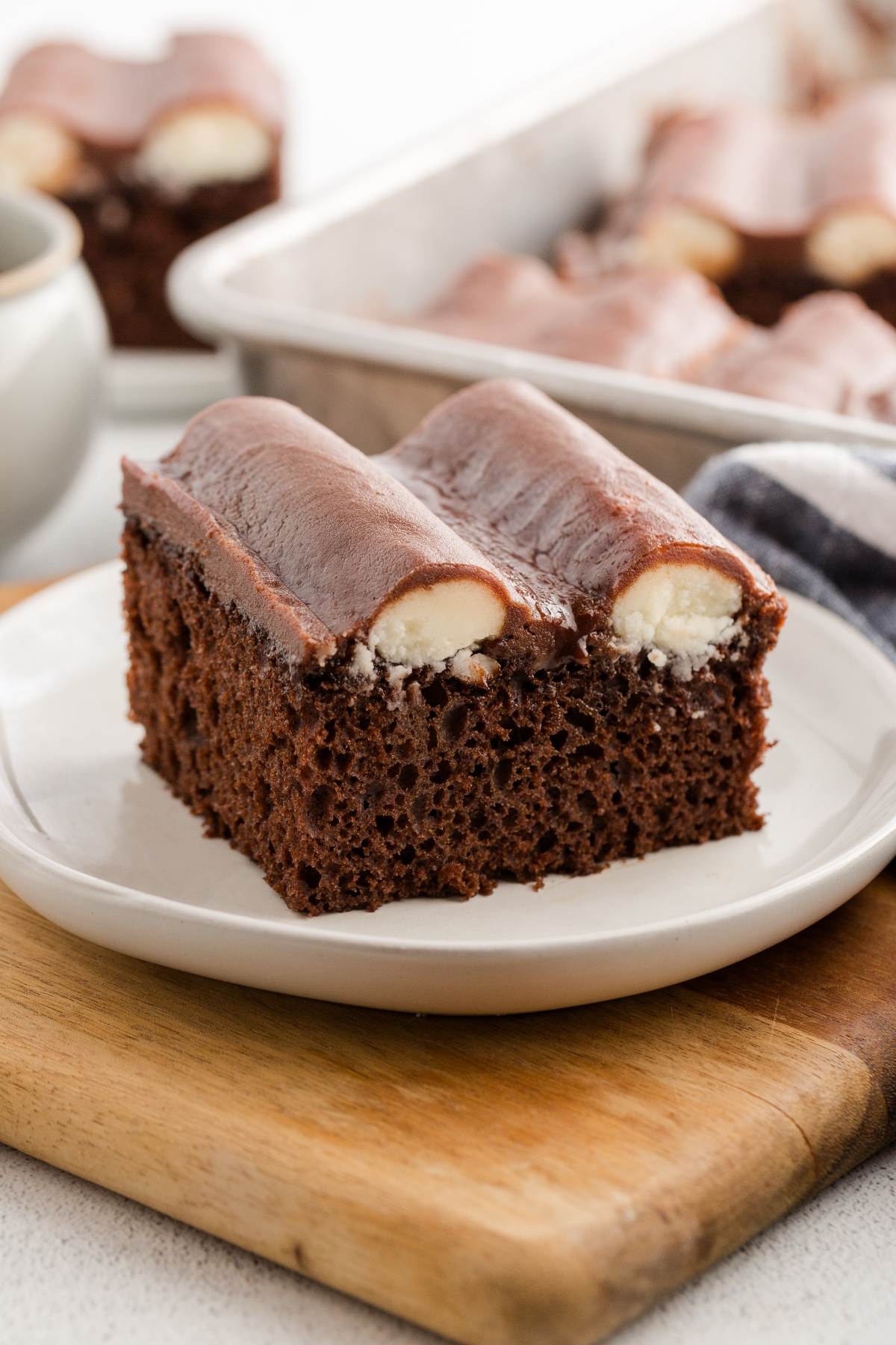 A slice of bumpy chocolate cake with cream filling on a white plate, with more cake in the background.