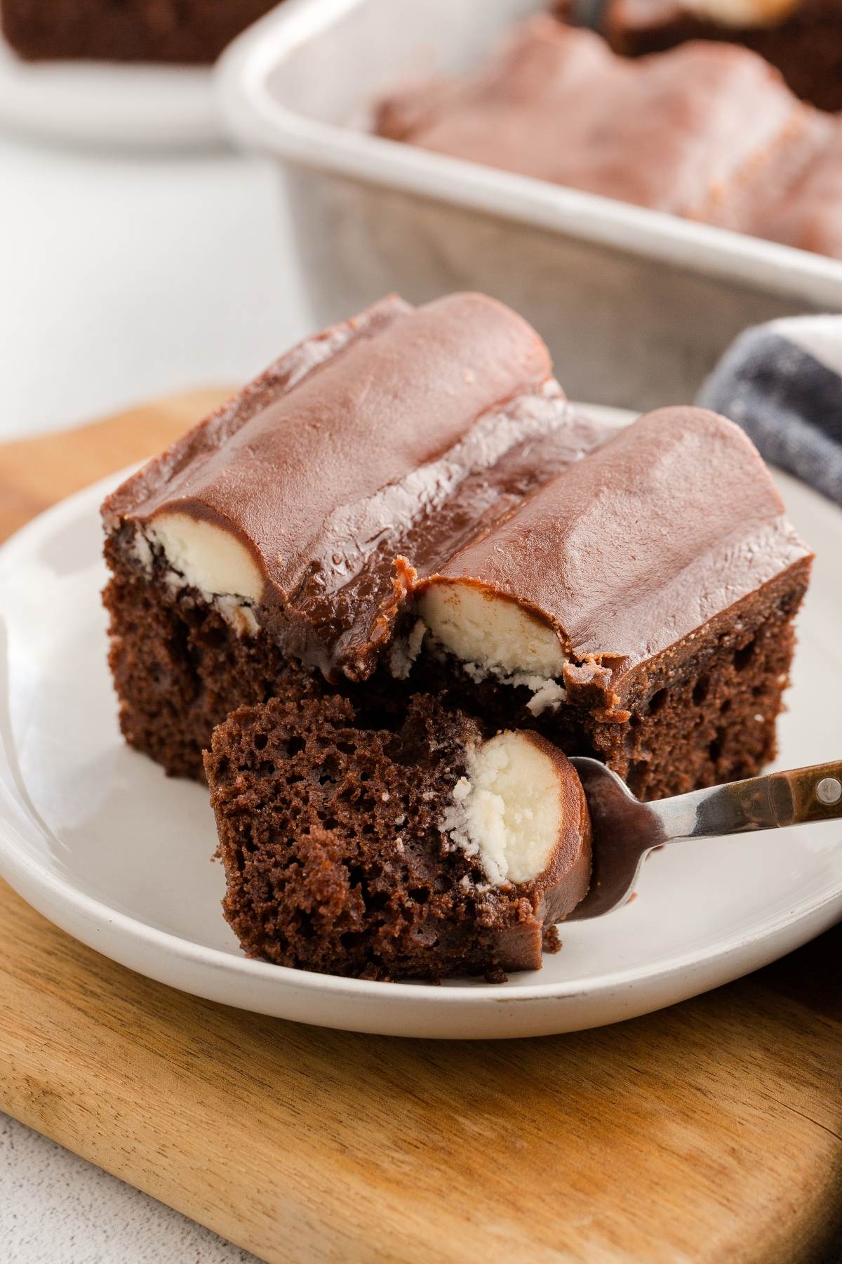A slice of chocolate cake with creamy filling and chocolate frosting on a white plate, with a forkful beside it.