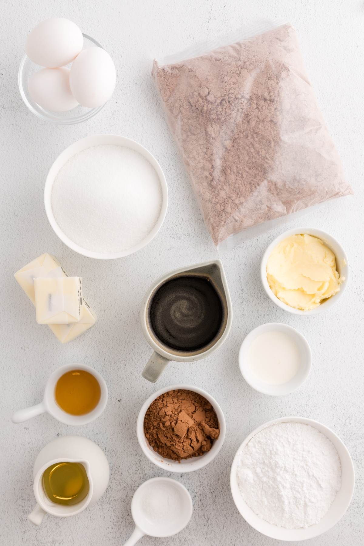 Assorted baking ingredients including eggs, sugar, butter, cocoa, oil, and flour arranged on a white surface.