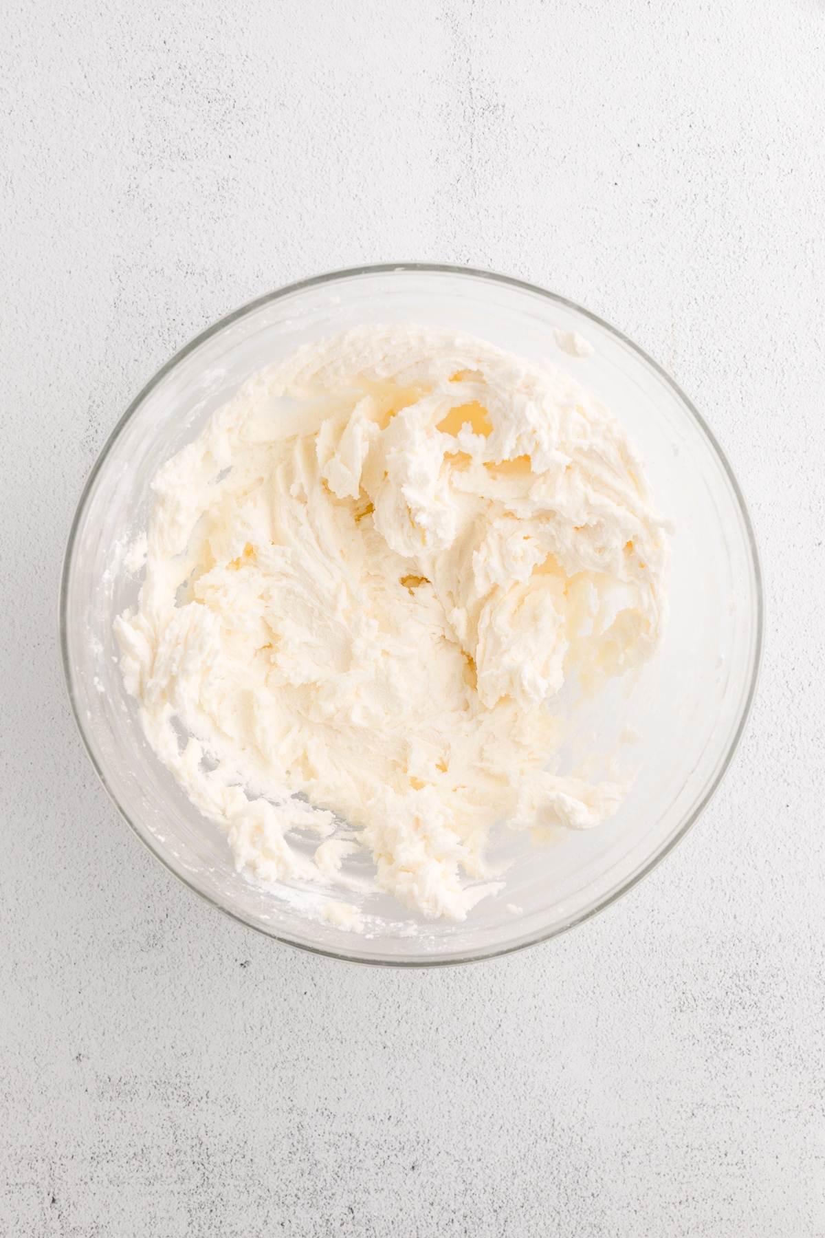 A glass bowl filled with whipped, creamy white frosting on a light textured surface.