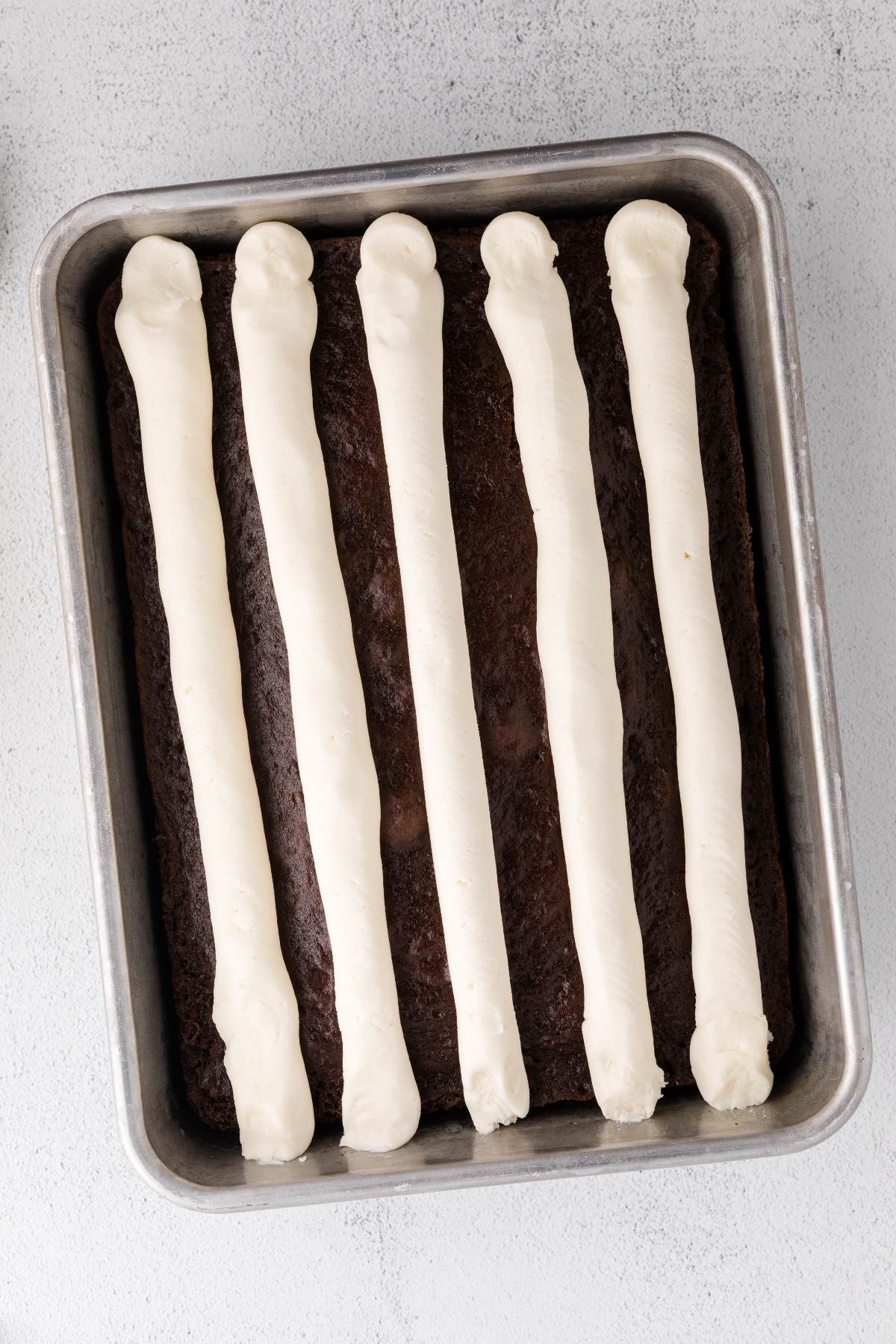 Rectangular chocolate cake topped with six evenly spaced vertical lines of white frosting in a metal pan.
