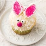 A bunny cupcake with pink ears, candy eyes, and a pink nose sits on a white plate—one of the cutest bunny cupcakes you'll ever see.
