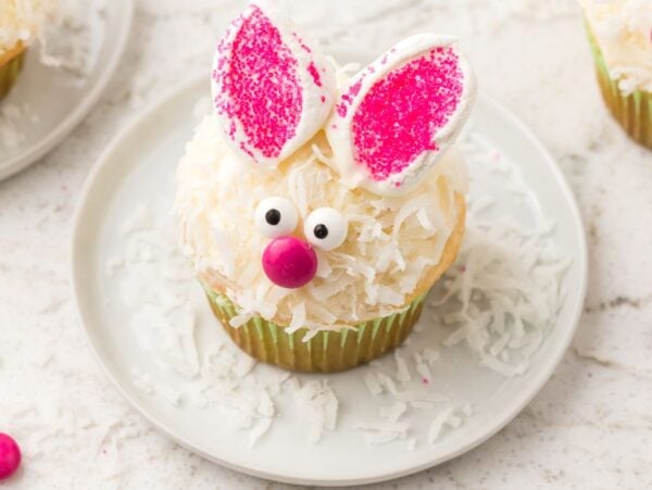 A bunny cupcake with pink ears, candy eyes, and a pink nose sits on a white plate—one of the cutest bunny cupcakes you'll ever see.