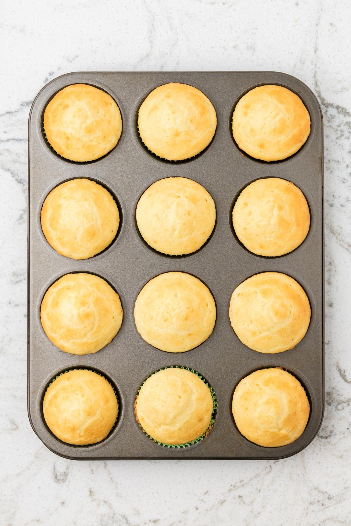 A muffin tray with twelve freshly baked golden muffins on a white marble countertop.