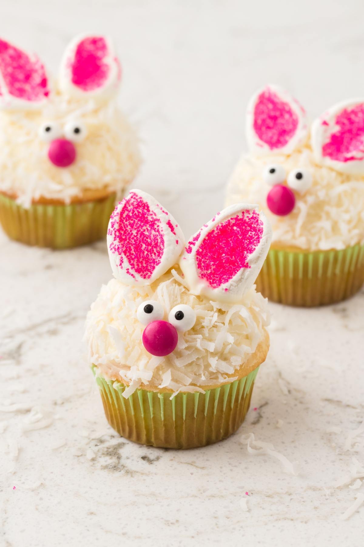 Three cupcakes decorated as bunnies with marshmallow ears, candy eyes, and pink noses on a white surface.