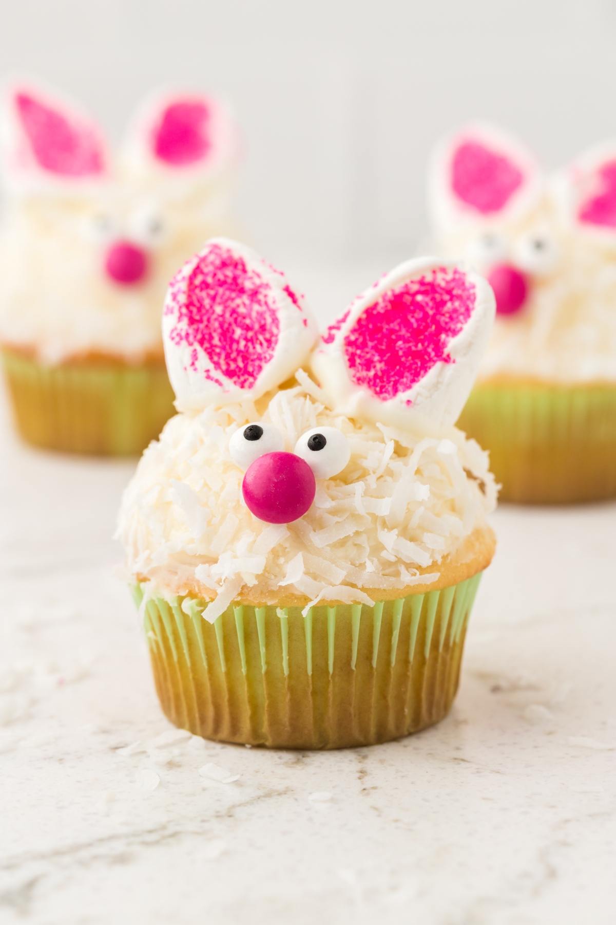 Cupcakes decorated as bunnies with pink ears, candy eyes, and coconut fur on a light surface.