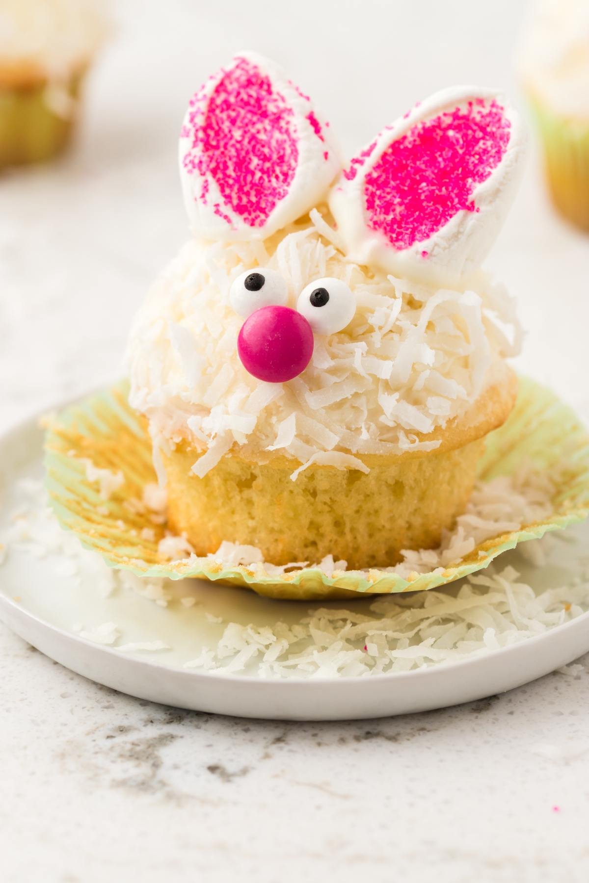 Cupcake decorated as a bunny with candy eyes, a pink nose, and marshmallow ears, on a white plate.