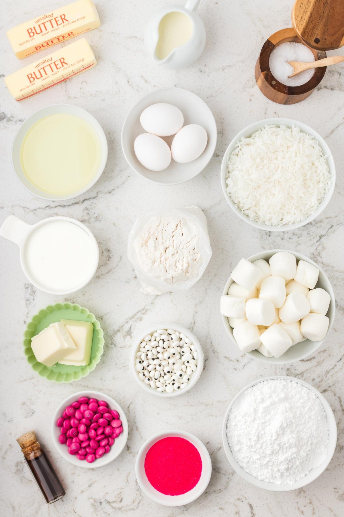Baking ingredients including butter, eggs, marshmallows, coconut, flour, sugar, candies, and milk on a marble surface.