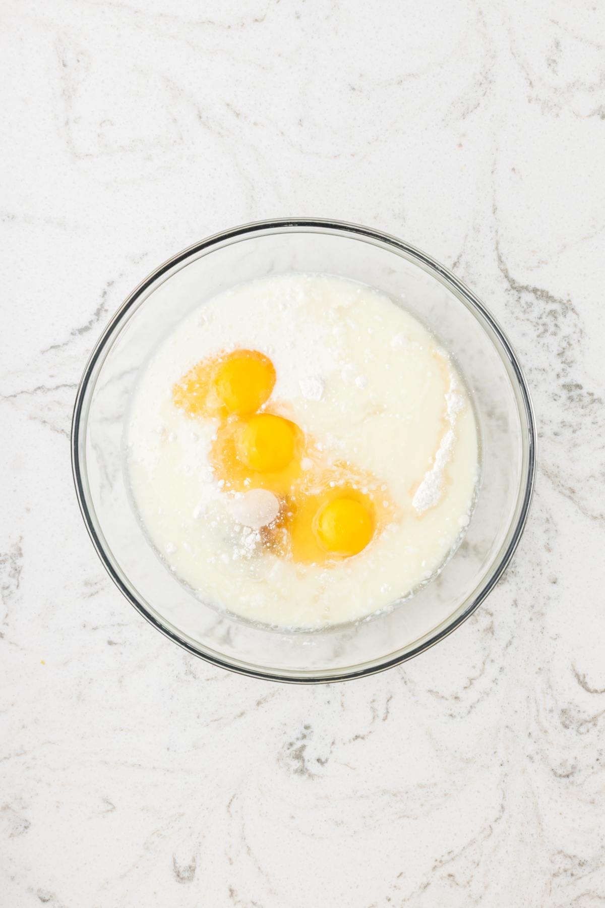 A glass bowl with three eggs, milk, and flour on a white marble countertop.