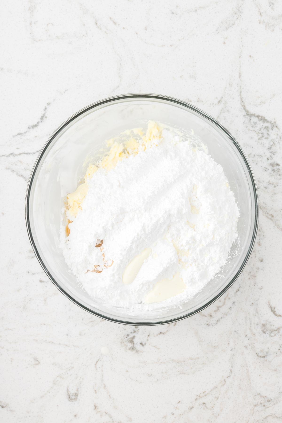 A glass bowl with butter and powdered sugar on a white marble countertop, ready to be mixed.