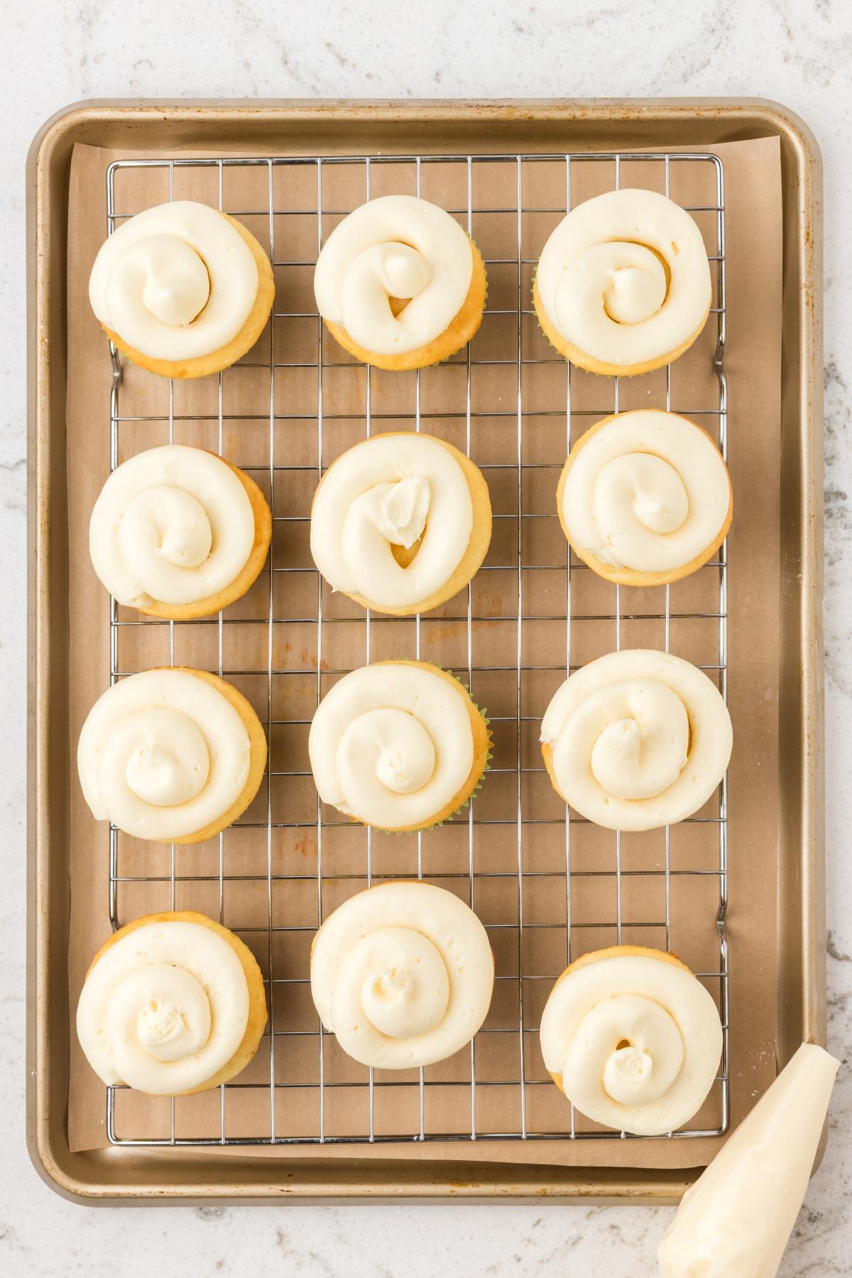 Twelve frosted cupcakes on a cooling rack set over a baking sheet, with a piping bag on the side.